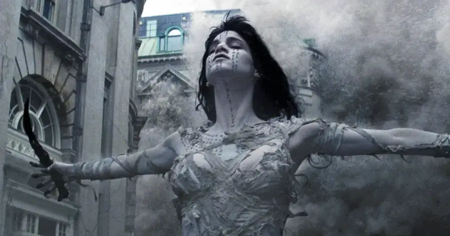 A woman wrapped in tattered, ancient bandages stands with arms outstretched, eyes closed, and head tilted back as dust and debris swirl dramatically around her in an urban setting.