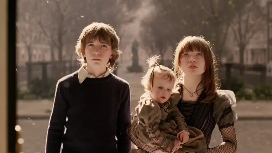 Three children stand outside looking up: an older boy in a dark sweater, a girl in a vintage dress holding a baby, with a sunlit, tree-lined street blurred in the background.