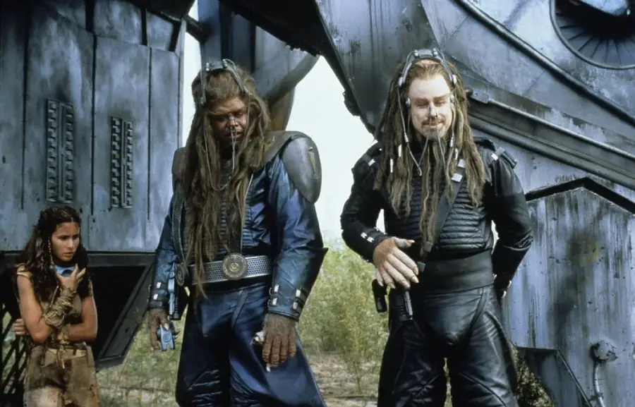 Three characters stand outside a large metallic structure; two tall figures wear dark futuristic armor and have long dreadlocks, while a woman in tattered clothing stands to the side, appearing cautious.