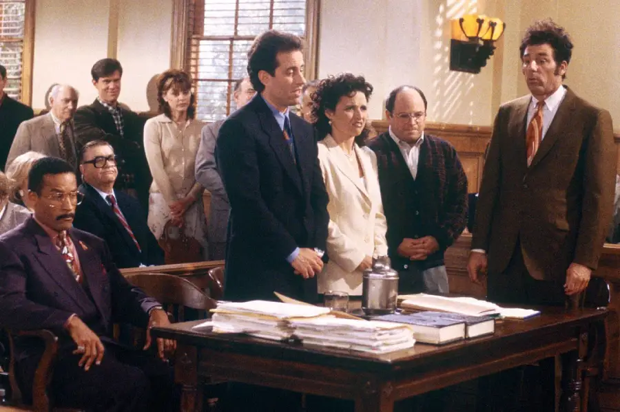 A courtroom scene with four people standing in front of a judge’s bench, with others seated and watching. The setting appears serious, with legal documents on the table and people dressed in formal and business attire.