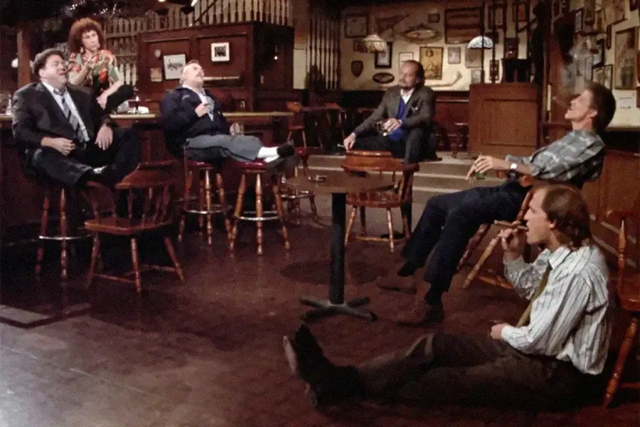 Six people relax in an empty bar. Four people sit on chairs around tables, one sits on the floor smoking, and one stands behind the bar. The scene has a cozy, dimly lit atmosphere with wooden decor.