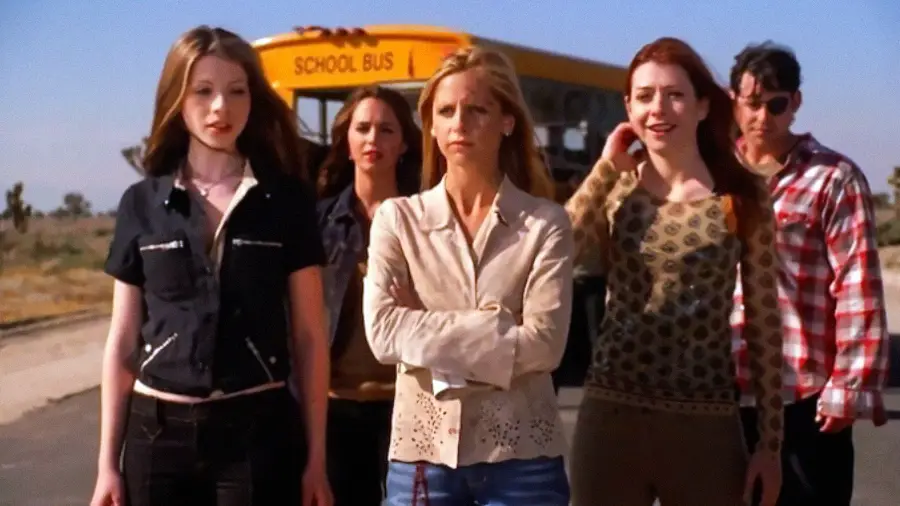 Five young people stand in front of a yellow school bus on a sunny day, looking serious and determined. The person in the center has arms crossed, while the rest stand close behind her. The background is outdoors with clear skies.