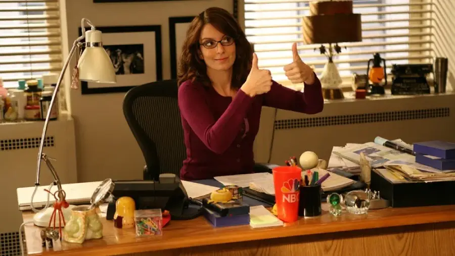 A woman with glasses and a maroon sweater sits at a cluttered office desk, smiling and giving two thumbs up. There are papers, office supplies, and an NBC mug on the desk, with framed photos on the wall behind her.