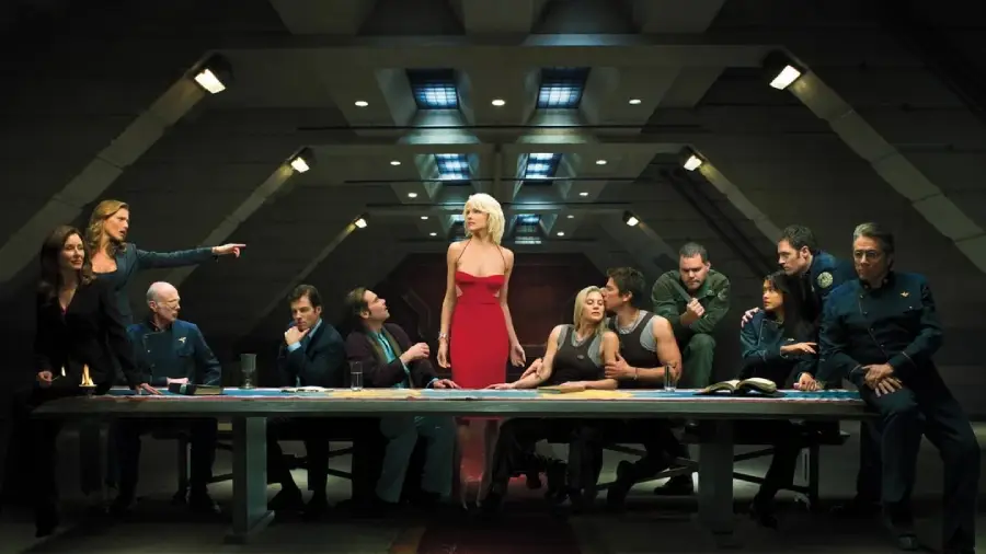 Twelve people sit or stand around a long table in a dim, industrial room. One woman in a red dress stands in the center, drawing attention. The group appears to be in a tense discussion, with dramatic lighting overhead.