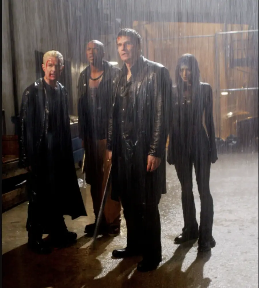 Four people stand together in the rain at night, all wearing dark clothing and looking ahead with serious expressions. The scene is dramatic, with rain pouring heavily around them in an urban setting.