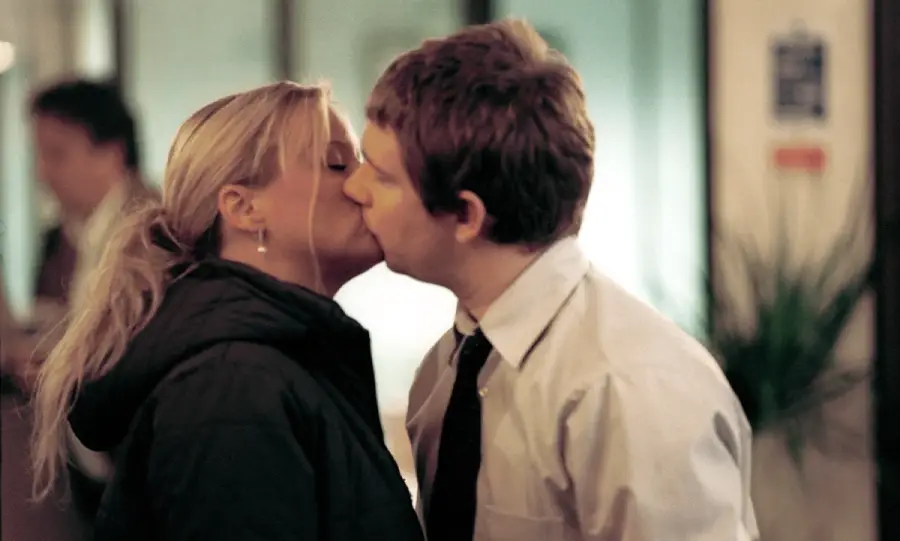 A woman in a dark jacket and a man in a white shirt and tie are kissing indoors with blurred people and plants in the background.