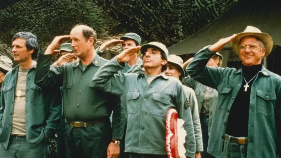 A group of men in military uniforms stand in a row outdoors saluting, some smiling. One man wears a cowboy hat and a cross necklace, while others have standard army caps. Trees and a tent are visible in the background.