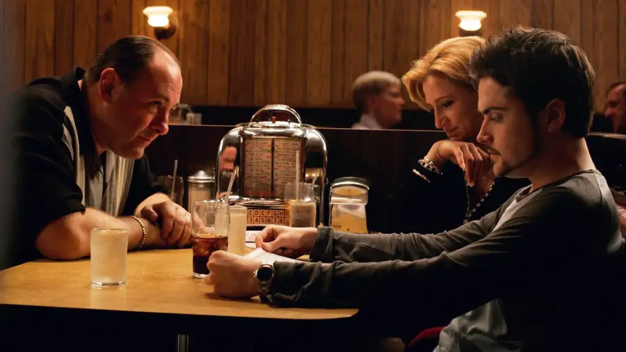 Three people sit closely at a dimly lit diner booth, two with drinks and one with folded hands. A jukebox sits on the table, and wood-paneled walls create a cozy, intimate atmosphere.