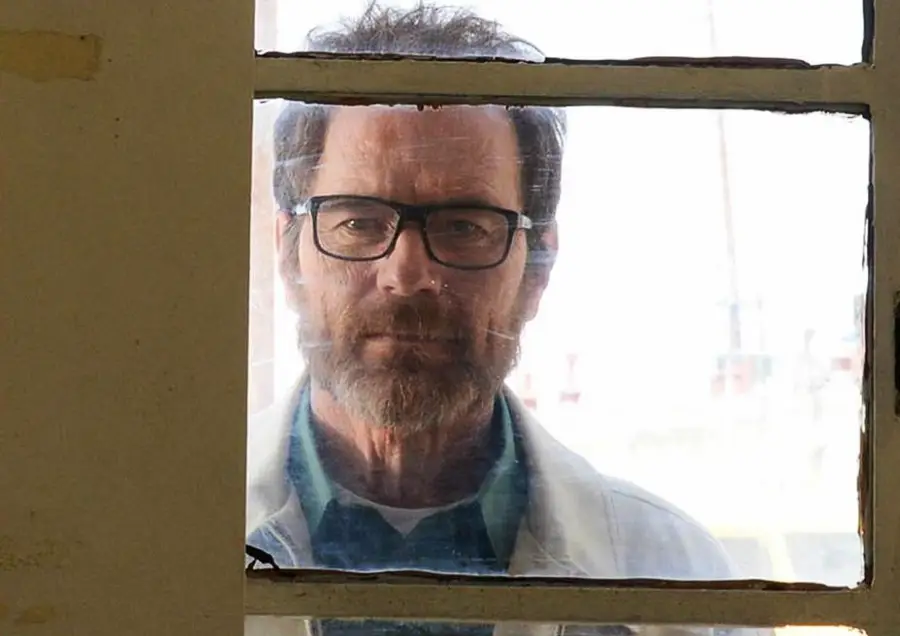 A bearded man with glasses and a serious expression looks through a window with white frames, seen from inside a building.