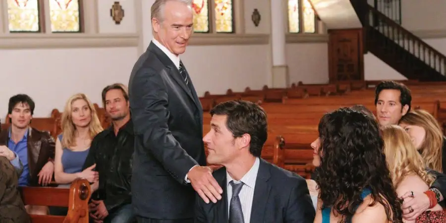 A group of people sit in wooden pews inside a church. An older man stands, resting his hand on the shoulder of a seated man in a suit, while others watch. Stained glass windows and a staircase are visible in the background.