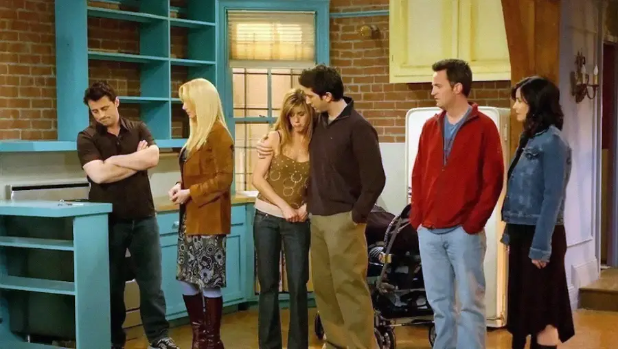 Six people stand in a nearly empty apartment, looking emotional. The room has teal shelves, a brick wall, and minimal furniture. Some hug or touch each other for comfort, suggesting a heartfelt farewell.