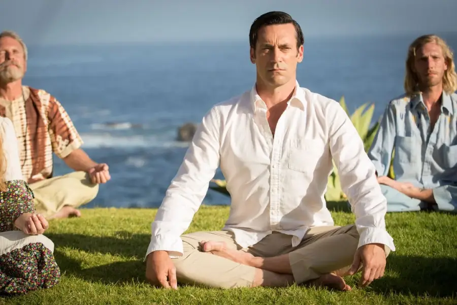 A man in a white shirt meditates cross-legged on grass, with two others beside him, against a backdrop of the ocean and blue sky.