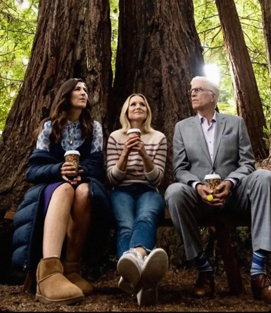 Three people sit on a bench in front of giant redwood trees, holding coffee cups. The person on the left wears a blue coat and boots, the center wears jeans and a striped sweater, and the right wears a gray suit.