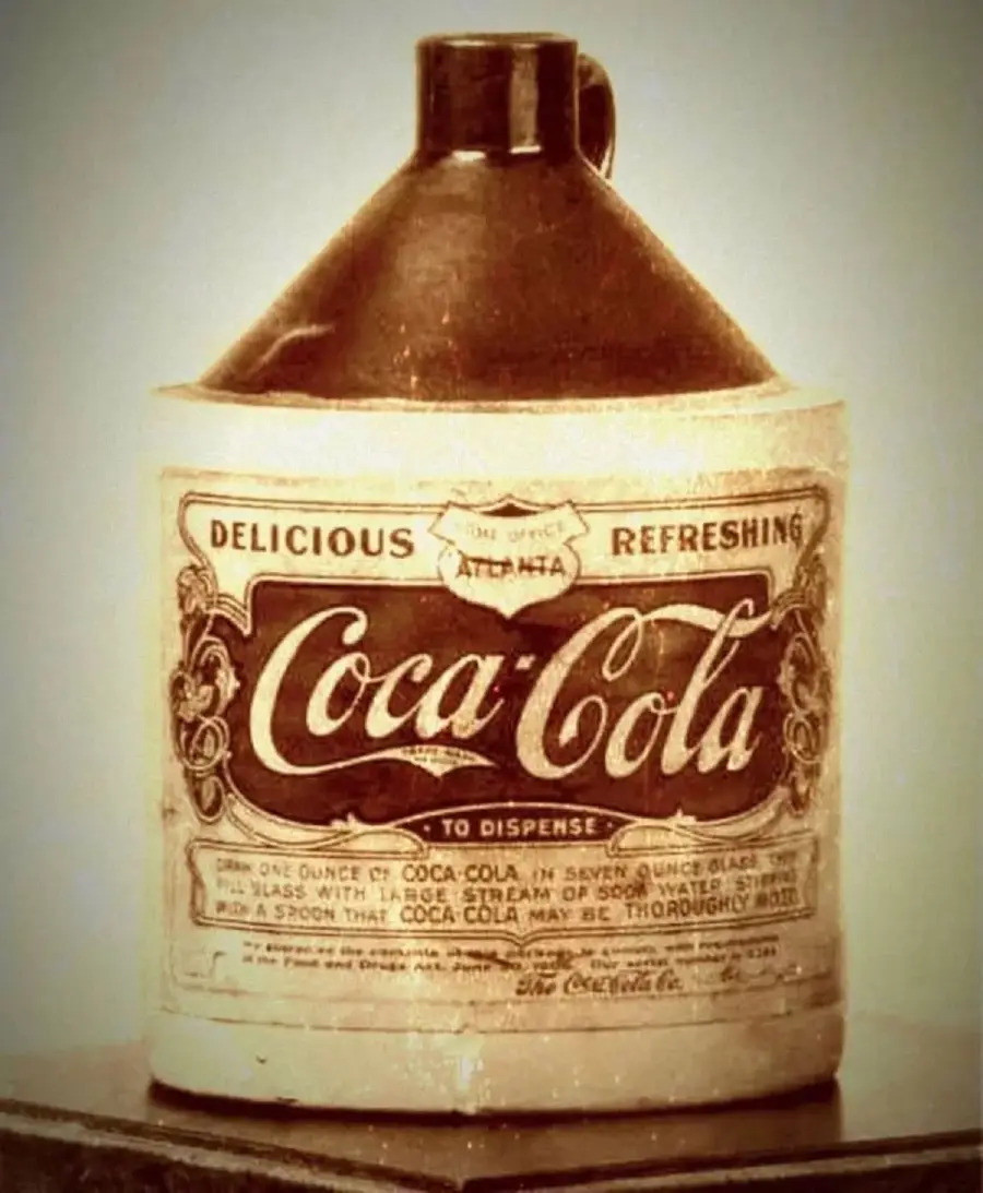 A vintage Coca-Cola syrup jug with an ornate label reading "Delicious Refreshing Coca-Cola," featuring mixing instructions and a dark brown handle on top. The jug looks old-fashioned and sepia-toned.