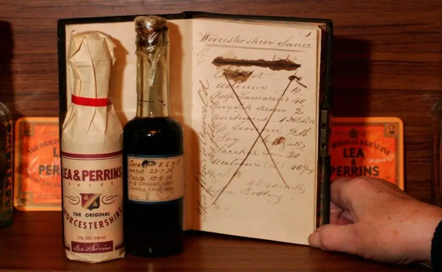 A hand holds open an old book displaying handwritten notes and strings, flanked by a bottle and a wrapped bottle of Lea & Perrins Worcestershire Sauce, with vintage labels in the background.