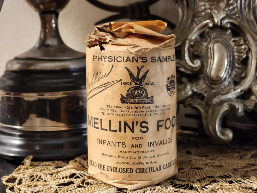 A vintage, cylindrical package labeled “Mellin’s Food for Infants and Invalids,” with aged, crumpled paper and handwritten notes, sits on a lace doily beside an ornate lamp and metallic decor.