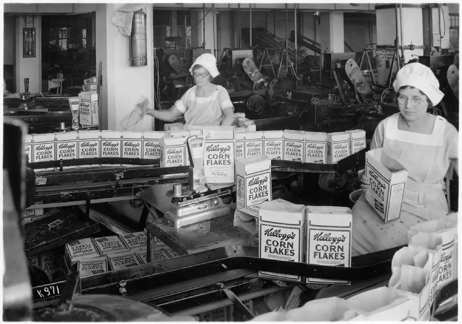 Two women in uniforms and caps pack Kellogg's Corn Flakes boxes on a production line in a factory filled with machinery. Stacks of cereal boxes are neatly arranged on the conveyor belt.