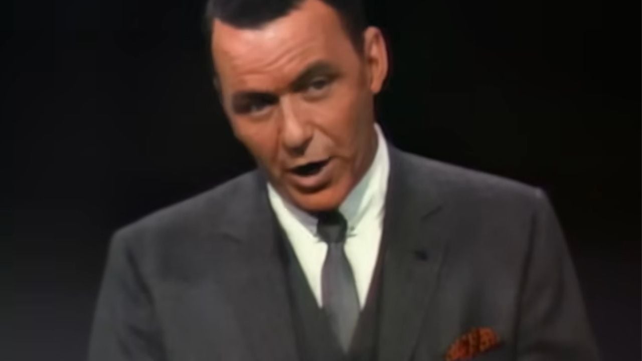 A man with dark hair wearing a gray suit, tie, and white shirt, stands against a dark background, speaking or singing.
