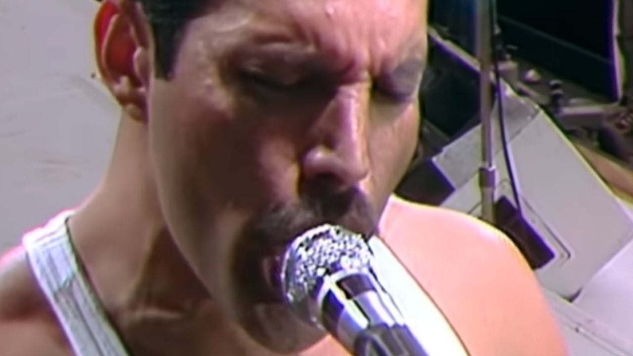 A close-up of a person with short dark hair and a mustache passionately singing into a sparkly microphone on stage, wearing a white tank top.