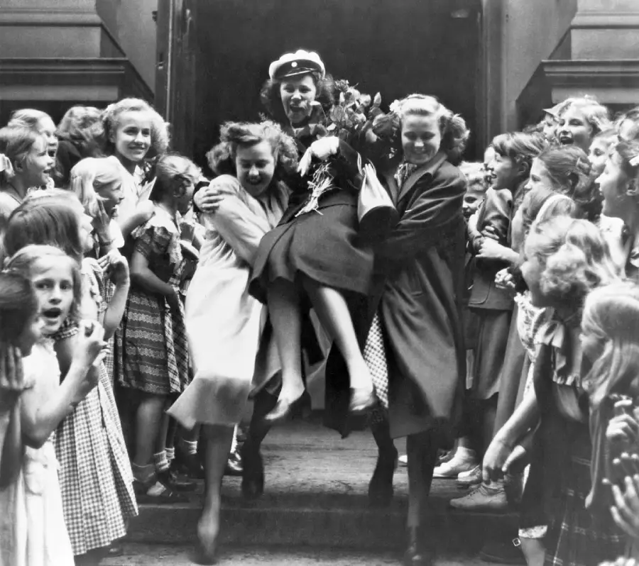 A group of smiling girls carry a friend while other girls cheer around them, celebrating on the steps of a building in this black and white vintage photo.