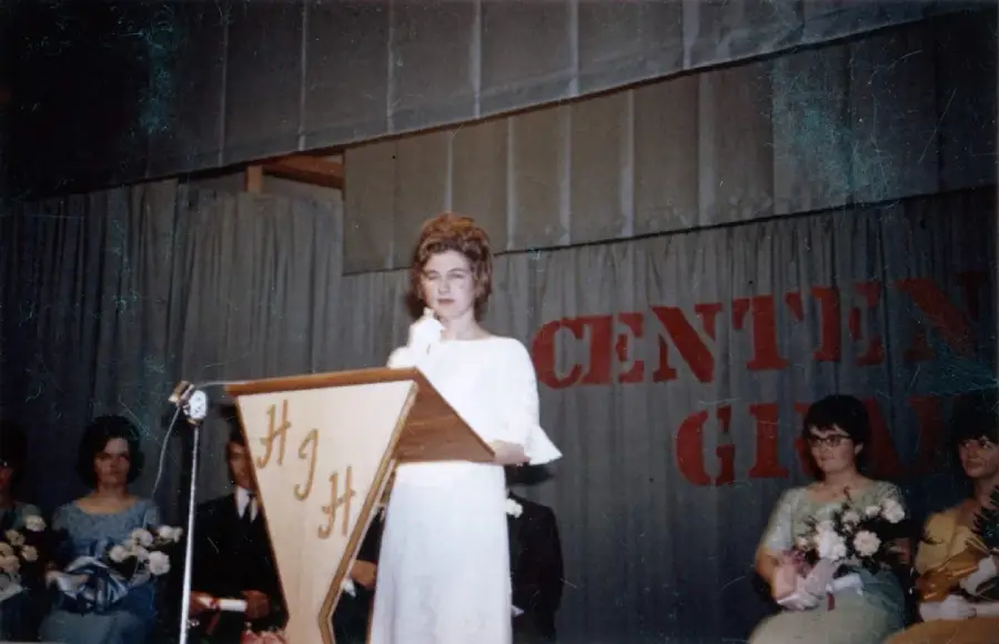 A young woman in a white dress stands at a wooden podium on a stage, speaking into a microphone. Several people are seated behind her, and large red letters on the gray curtain spell "CENTEN" and "GRAD.