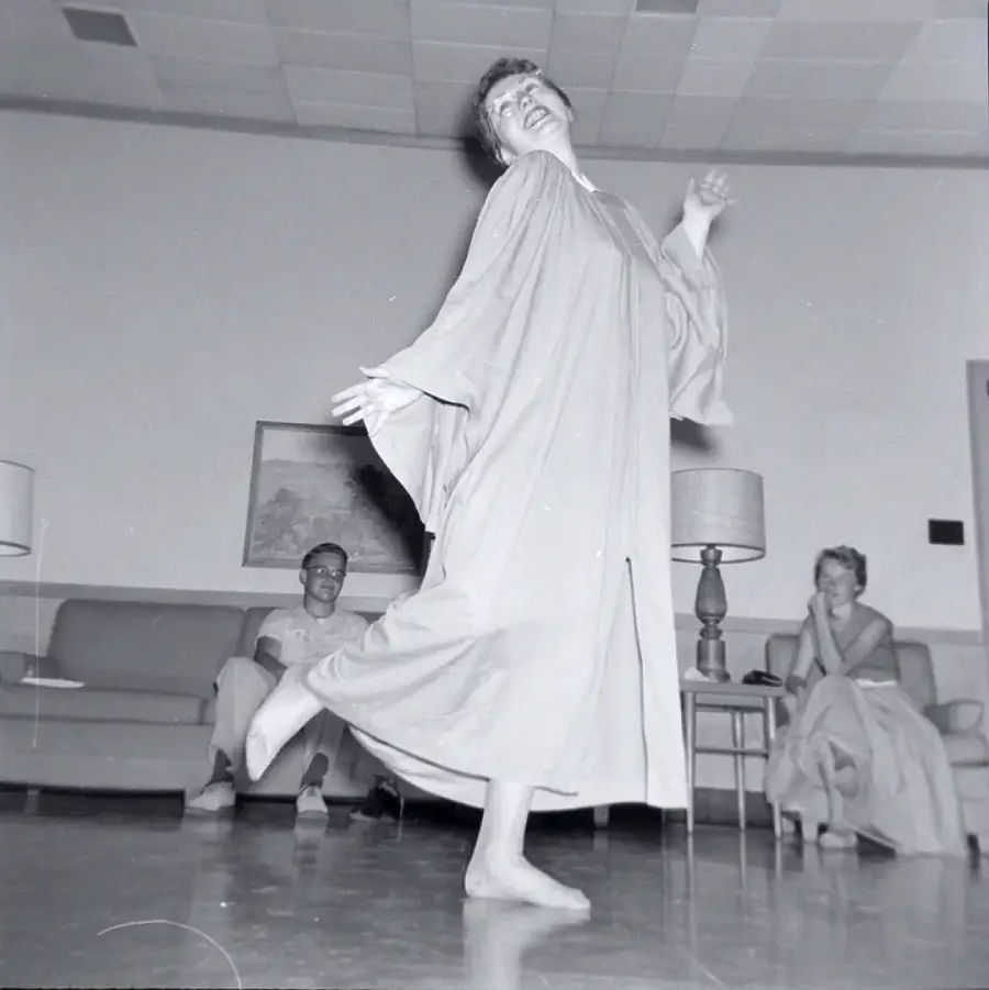 A barefoot woman in a loose dress joyfully dances in a living room while two people sitting on a couch watch her, one smiling and the other clapping. The room has lamps, a sofa, and a painting on the wall.