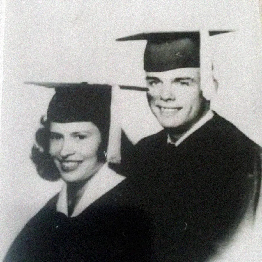 Black and white photo of two smiling people wearing graduation caps and gowns, posing closely together, likely on their graduation day.