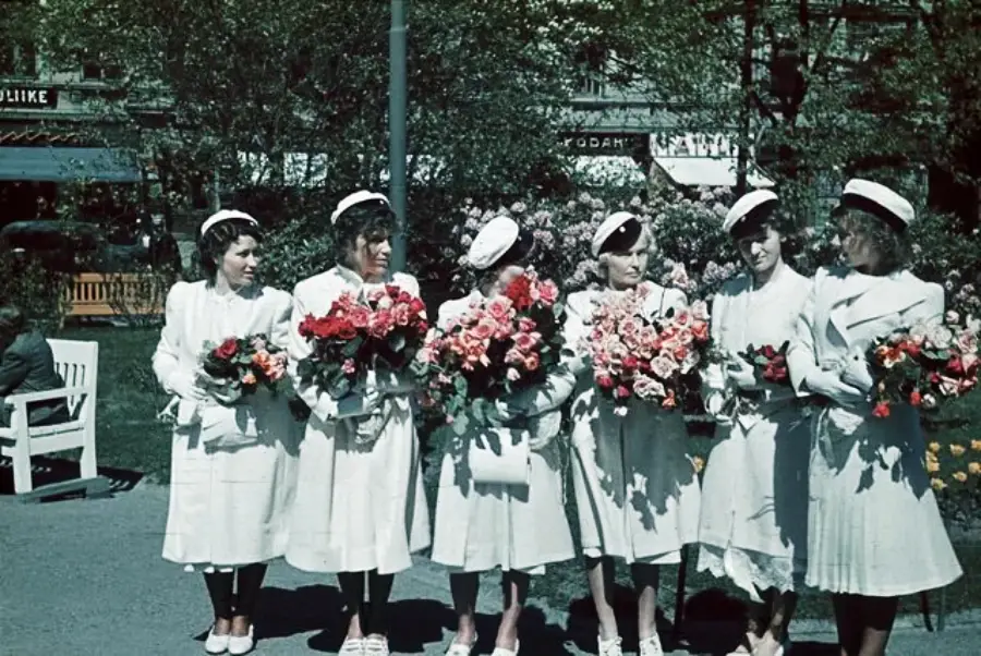 Six women dressed in white outfits and hats stand outdoors in a park, each holding large bouquets of colorful flowers. Trees and buildings are visible in the background.