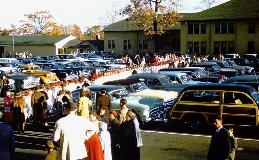 A large crowd gathers in a parking lot filled with vintage cars, watching a parade of people in red and white uniforms walk in a line. Trees with autumn leaves and buildings are visible in the background.