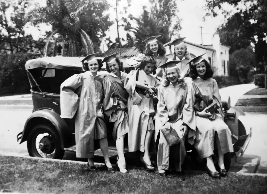 Seven graduates in caps and gowns smile and pose together on the back of a vintage car parked on a suburban street, with trees and houses in the background.