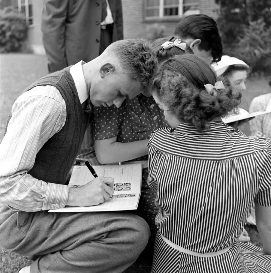 A young man writes on a clipboard while sitting outside with a group of people. They are closely gathered, and most are wearing patterned clothing. The scene appears candid and from a past era.