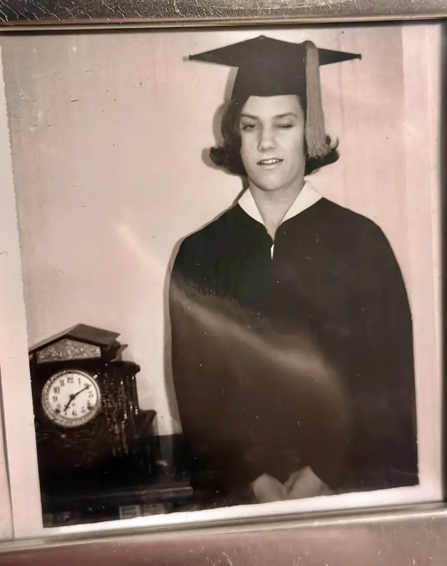 Black and white photo of a person in a graduation cap and gown standing indoors, hands folded. There is an ornate clock showing 12:02 on a table to the left of the person.