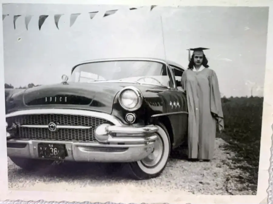 A young woman in a graduation gown and cap stands beside a vintage Buick car on a gravel road. Triangular flags are strung above, and the scene appears to be from the mid-20th century.
