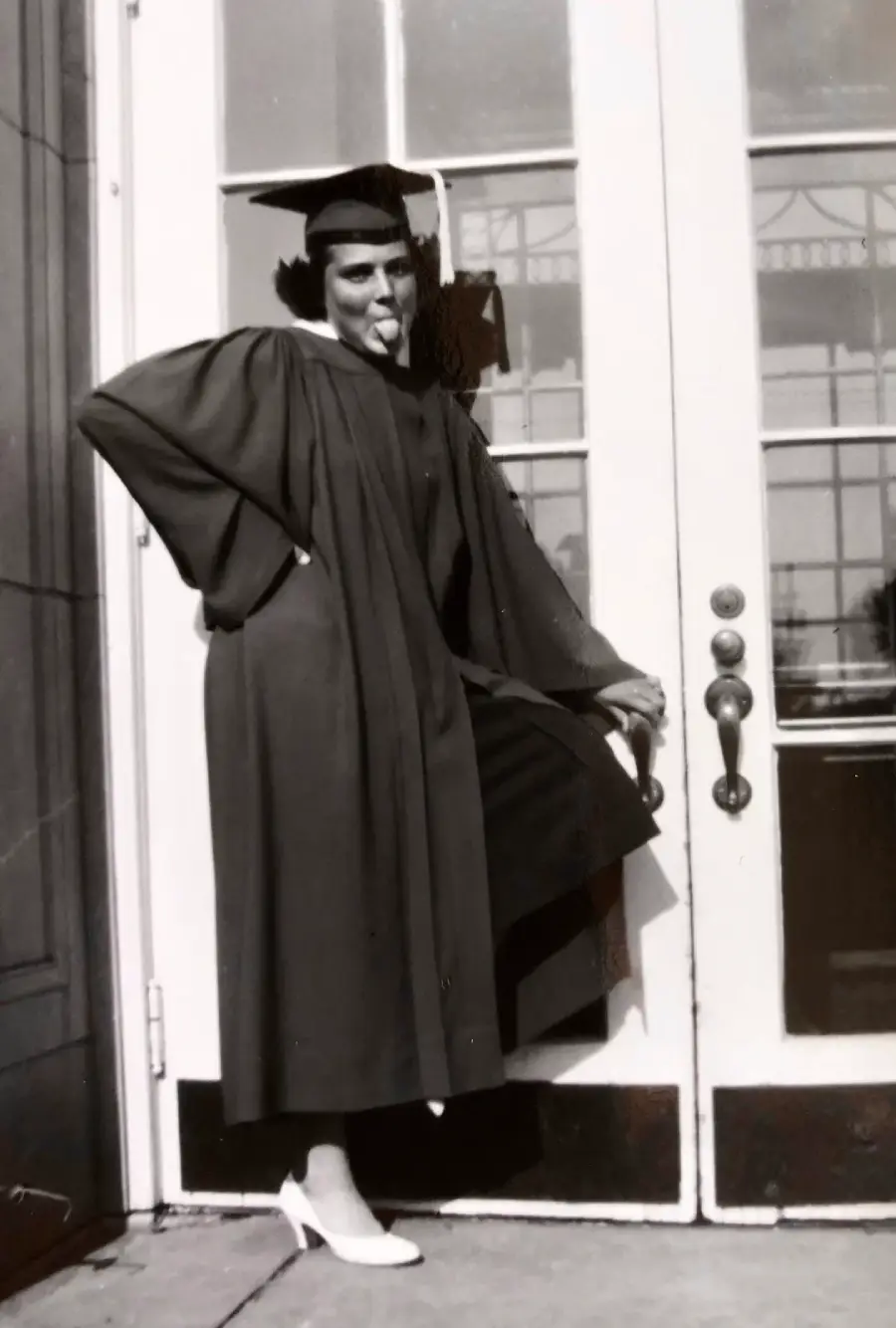 A person in a graduation cap and gown poses playfully, sticking out their tongue and lifting one leg while standing in front of glass double doors.