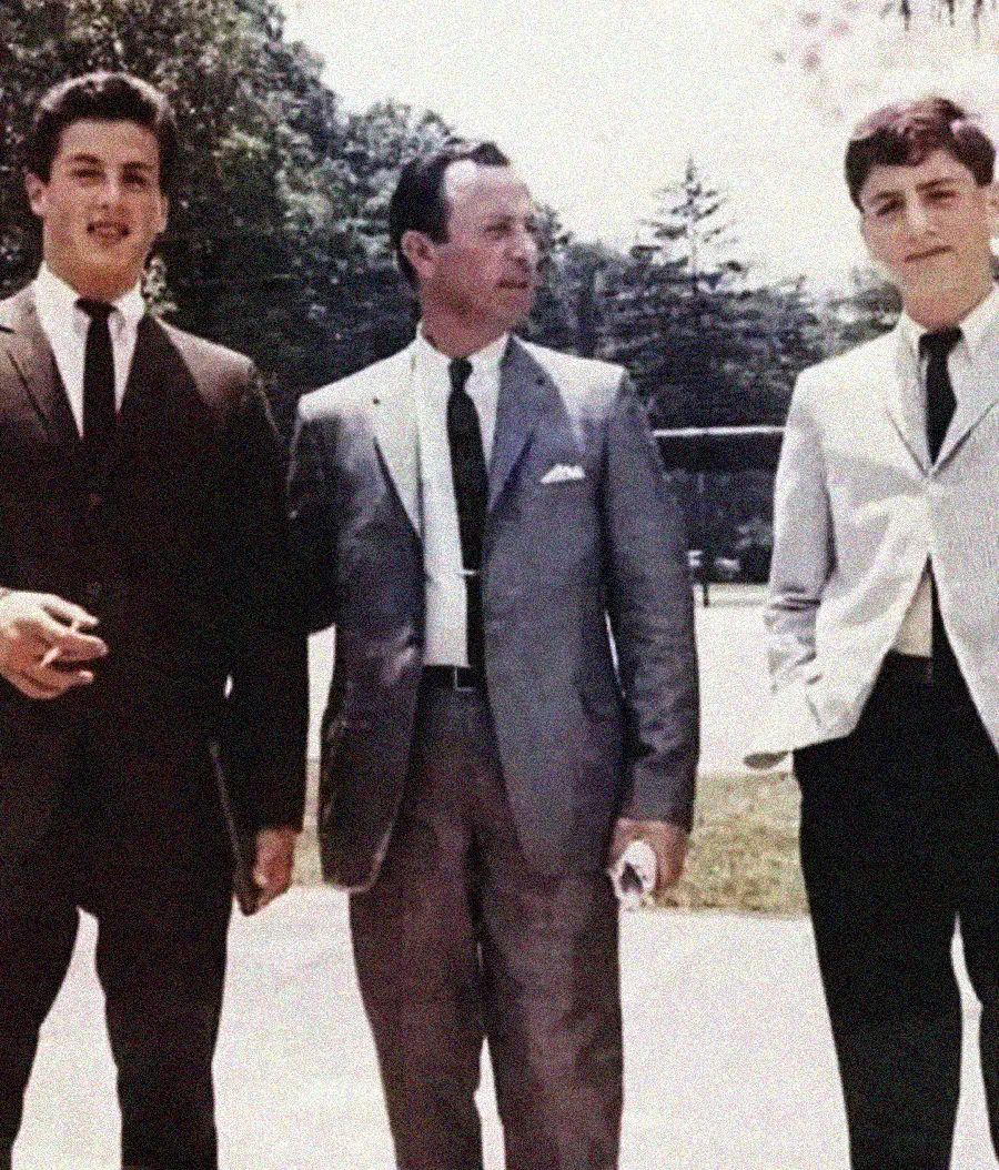 Three men stand outdoors in formal suits, posing for a photo. The man in the center wears a gray suit, while the men on either side wear dark and light suits. Trees and greenery are visible in the background.