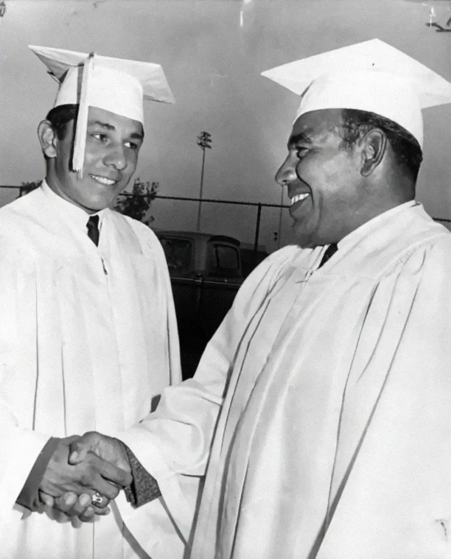Two men wearing graduation caps and gowns smile and shake hands outdoors, celebrating their graduation. A vintage car and sports field can be seen in the background.