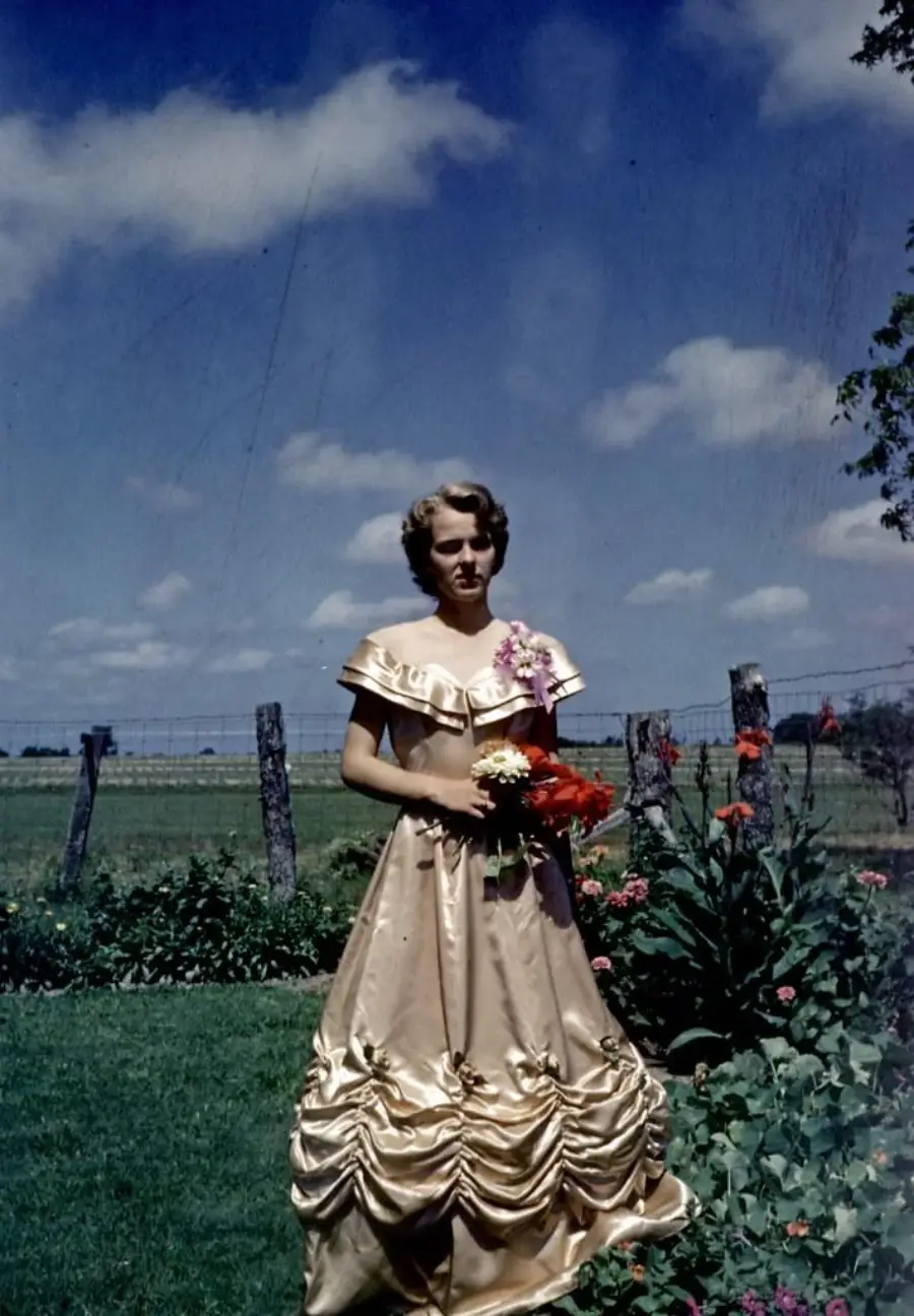 A woman in a shiny, formal gown stands on grass with a bouquet of flowers, in front of a garden and wire fence under a blue sky with scattered clouds. Fields and trees are visible in the background.