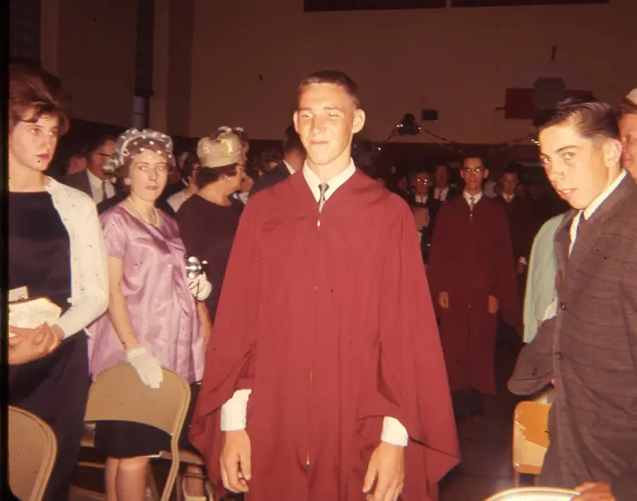 A young man in a maroon graduation gown stands in the center of a crowded indoor ceremony, surrounded by other students in suits and women in formal attire, hats, and gloves.