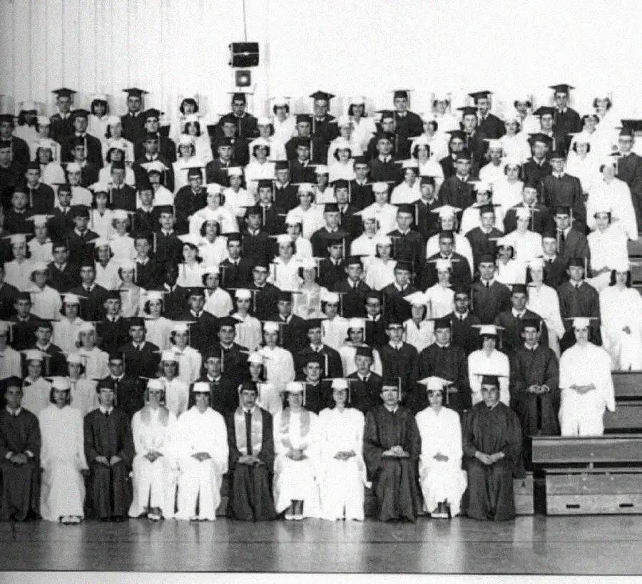 Black and white photo of a large group of graduates in caps and gowns, arranged in several rows on bleachers inside a gymnasium, posing for a formal graduation portrait.