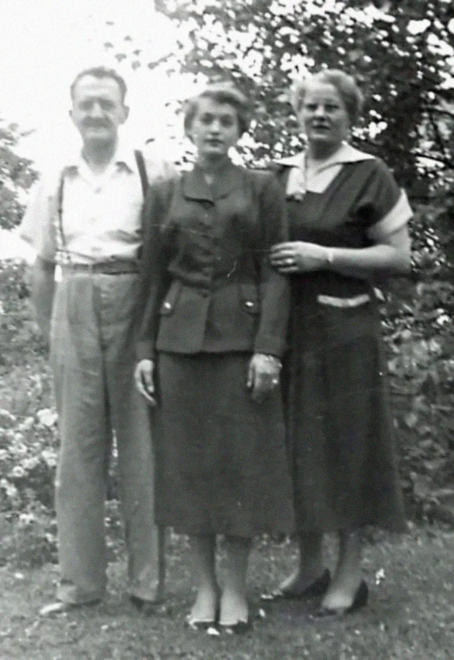 A black-and-white photo of three adults standing outdoors. An older man in suspenders stands to the left, a young woman in a skirt suit is in the center, and an older woman in a dress stands to the right. Trees are in the background.