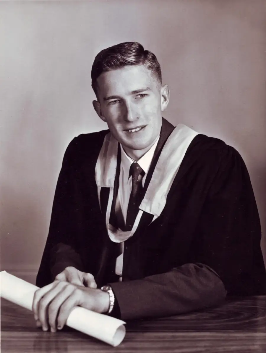 A young man in a graduation gown and tie sits at a table holding a rolled diploma, looking slightly to the side. The image is in black and white.