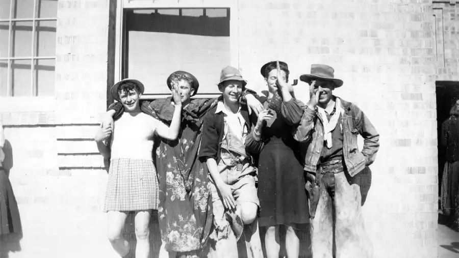 Five people stand in front of a brick building, smiling and posing playfully. Some are wearing dresses and hats, and a few have exaggerated makeup or costumes, suggesting a lighthearted or humorous moment.