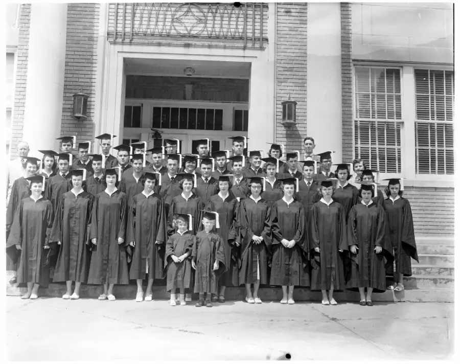 A large group of students wearing graduation caps and gowns stands in front of a brick school building, posing for a formal group photo. Two young children in caps and gowns stand in the front row.