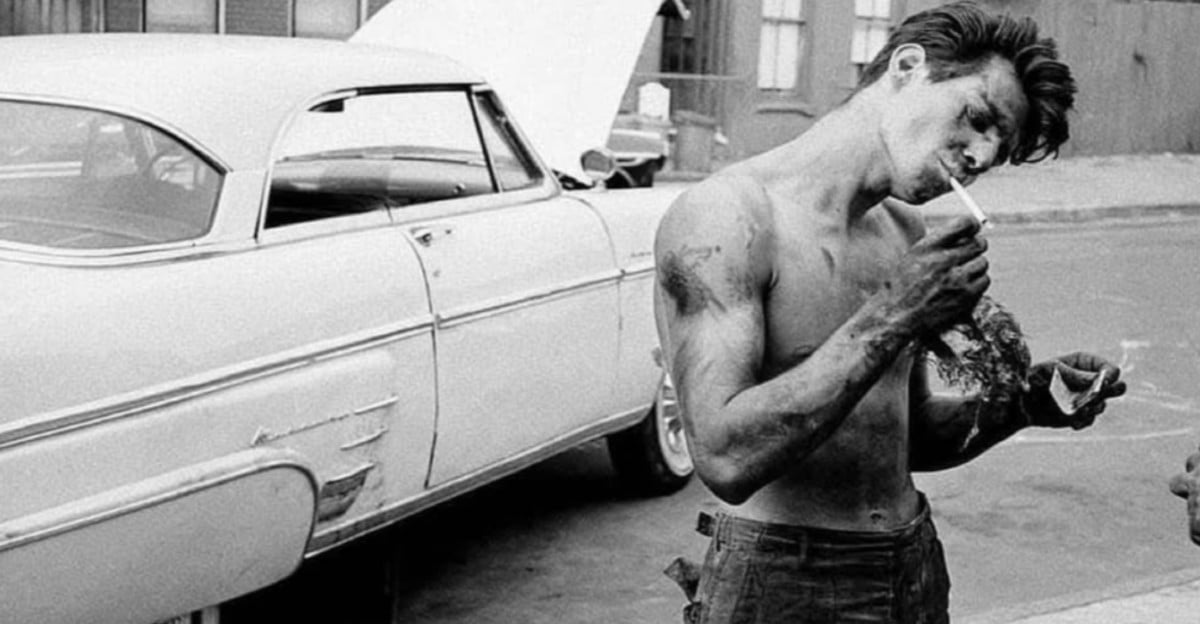 A shirtless man with dirty arms and face lights a cigarette while standing next to a vintage car with an open hood on a street.