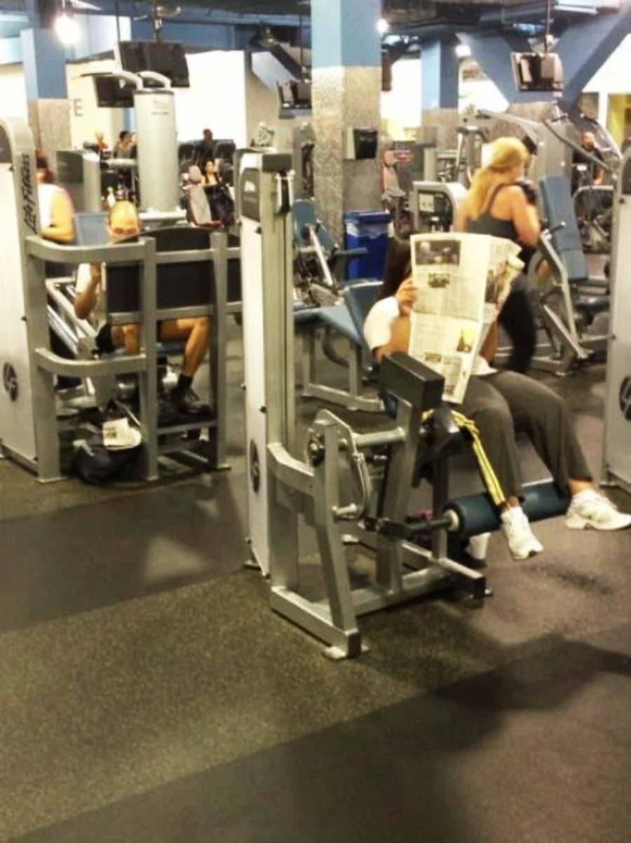 A person sits on a leg exercise machine at a gym, holding and reading a newspaper, while other people work out on various machines in the background.