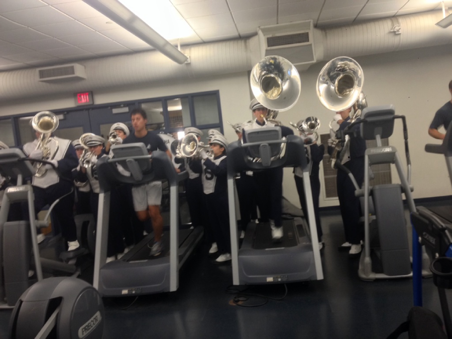 A marching band in uniform plays brass instruments while standing on treadmills in a gym, alongside a person in athletic clothes also using a treadmill.