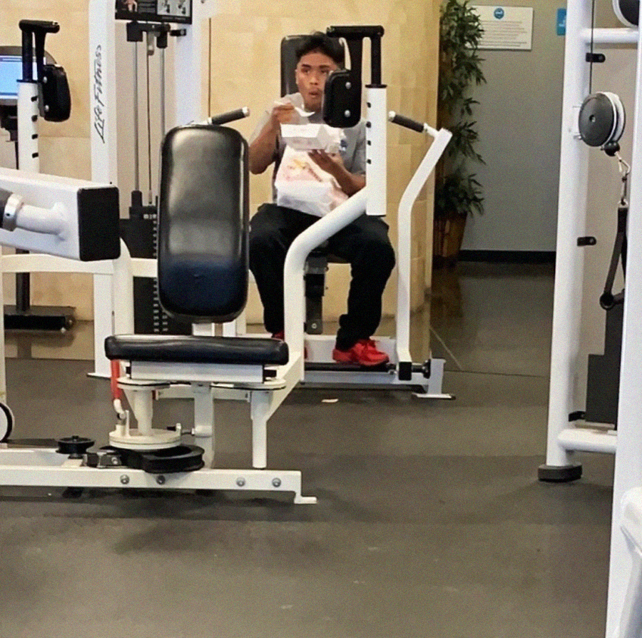 A man sits on a weight machine in a gym, eating from a takeout container with a fork. He is wearing black pants and red shoes, with gym equipment visible around him.