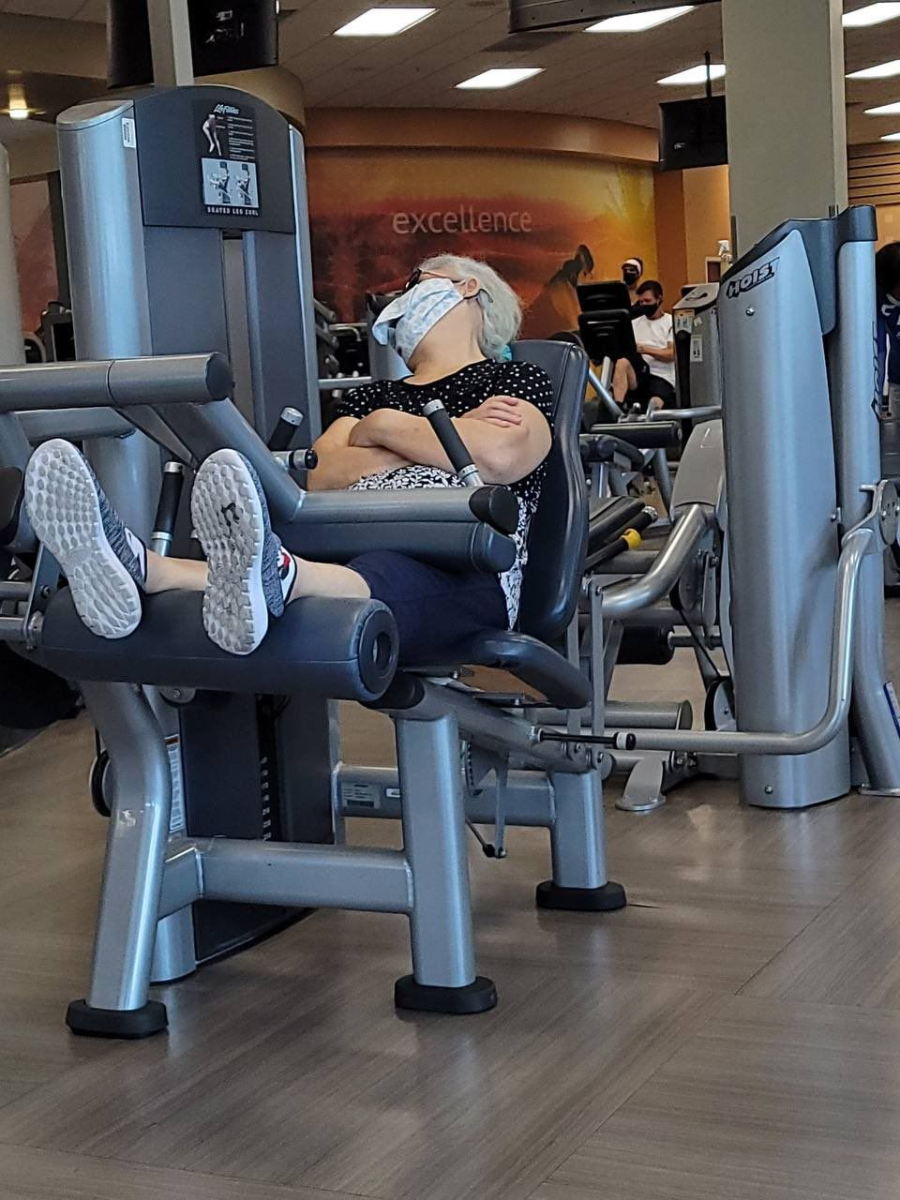 An older woman with a face mask sleeps with her arms and legs crossed on a leg extension machine in a gym. Other gym-goers are using equipment in the background.