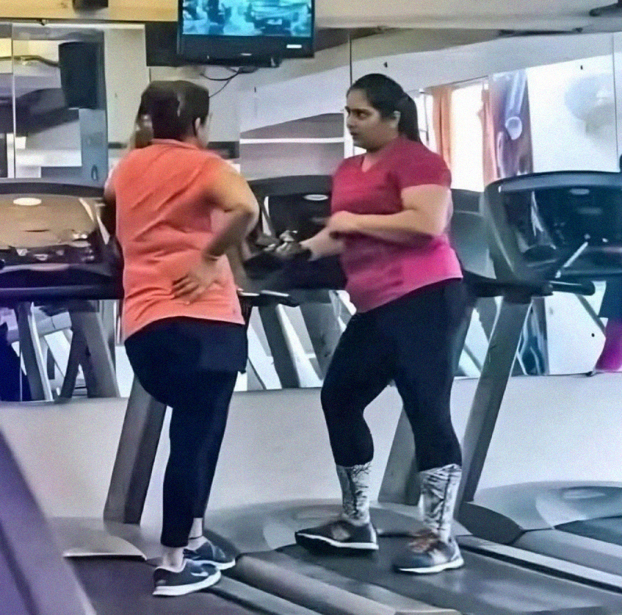 Two women are standing on treadmills at a gym, facing each other and talking. One has her hands on her hips, and the other is gesturing with her hands. Both are dressed in athletic wear.