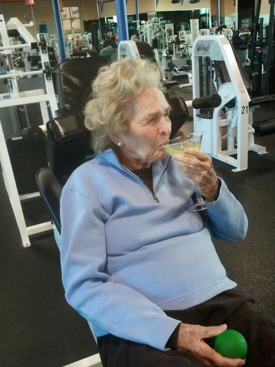 An elderly woman in a light blue sweater sits on exercise equipment at a gym, holding a green stress ball in one hand and sipping a cocktail from a martini glass. Gym machines and weights are visible in the background.